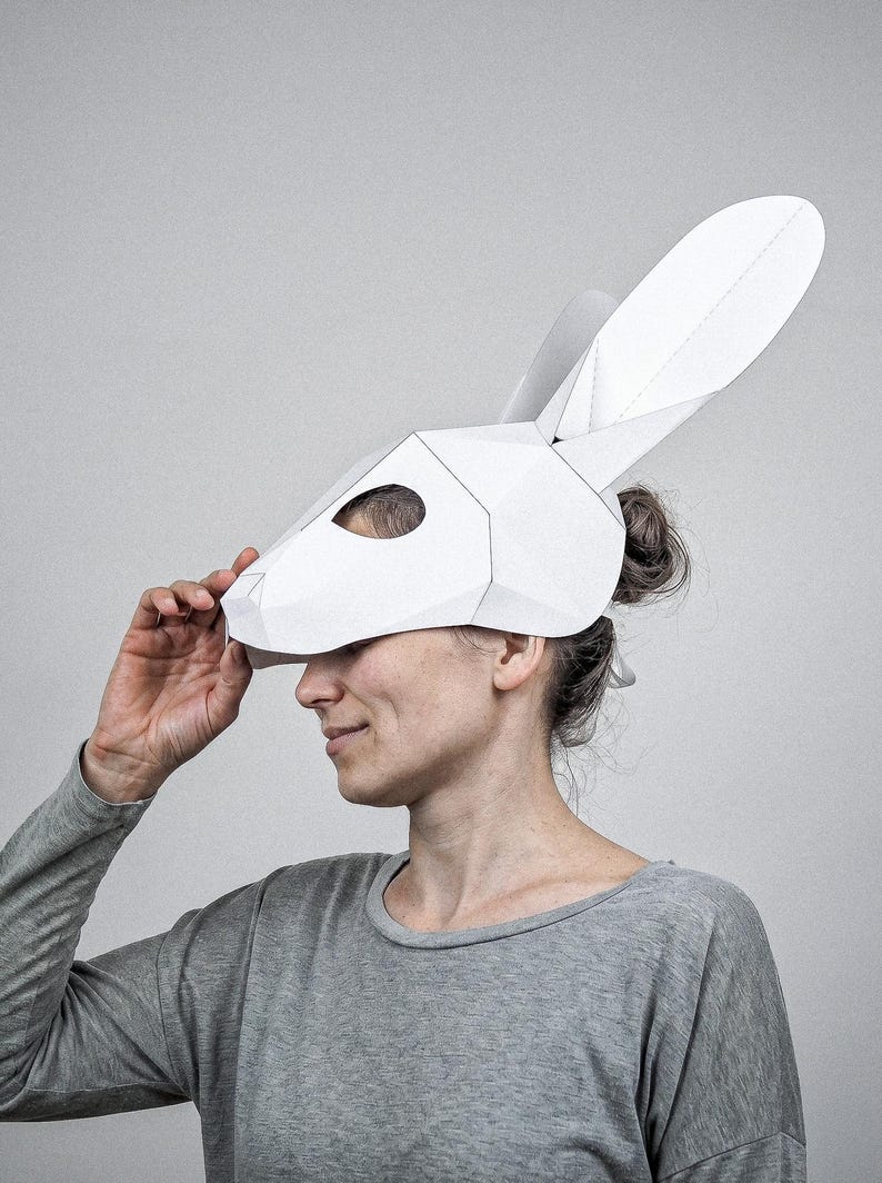 May include: A white, geometric rabbit mask with long ears, made from folded paper. A person wears the mask, paired with a grey long-sleeved top. The background is a plain, light grey.