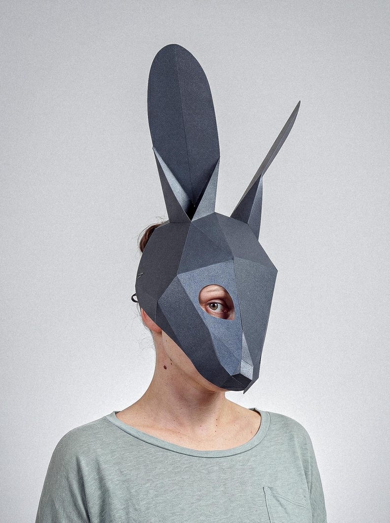 May include: A dark grey geometric rabbit mask with long ears and pointed features. The mask covers the face, with an eye visible through an oval opening. The person is wearing a light green top.