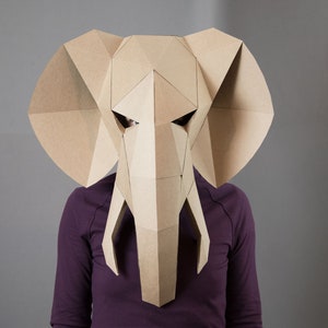 May include: A geometric elephant mask constructed from tan cardboard, featuring large flat ears, a central trunk, and two downward-pointing tusks. The mask is composed of multiple angular, faceted panels that form the shape of an elephant head.