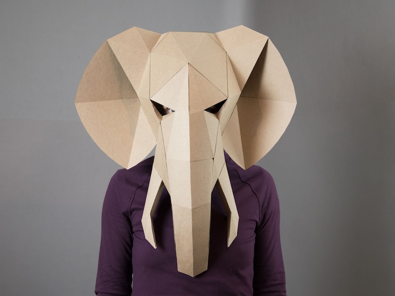 May include: A geometric elephant mask constructed from tan cardboard, featuring large flat ears, a central trunk, and two downward-pointing tusks. The mask is composed of multiple angular, faceted panels that form the shape of an elephant head.