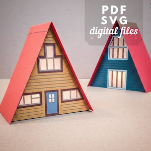 May include: Two papercraft A-frame houses with red roofs and detailed facades. One house is tan with a blue door, the other is blue. The image includes the text "PDF SVG digital files".