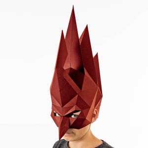 K&ouml;nnte beinhalten: Eine geometrische, burgunderfarbene Maske mit scharfen, kantigen Merkmalen. Die Maske bedeckt das Gesicht und erstreckt sich nach oben, &auml;hnlich einer Krone oder Kopfbedeckung. Das Design besteht aus gefaltetem Papier oder Karton und erzeugt einen auff&auml;lligen, dreidimensionalen Effekt.