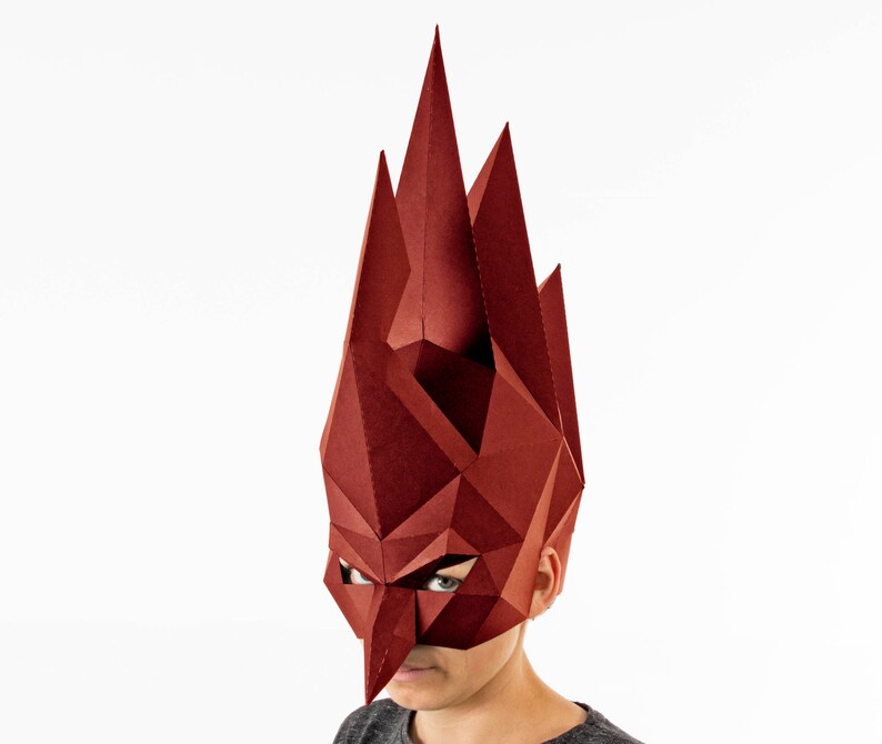K&ouml;nnte beinhalten: Eine geometrische, burgunderfarbene Maske mit scharfen, kantigen Merkmalen. Die Maske bedeckt das Gesicht und erstreckt sich nach oben, &auml;hnlich einer Krone oder Kopfbedeckung. Das Design besteht aus gefaltetem Papier oder Karton und erzeugt einen auff&auml;lligen, dreidimensionalen Effekt.