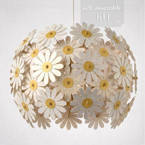 May include: A spherical pendant light fixture constructed from multiple white daisy-shaped pieces with yellow centers. The design features a layered floral pattern. A label in the upper right corner displays the text self assembly KIT.