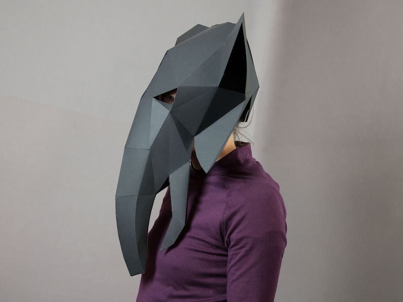 May include: A dark gray, low-poly paper mask shaped like an elephant head. The mask features a geometric, faceted design with a long trunk, pointed ears, and triangular eye openings, constructed from multiple angular paper panels.