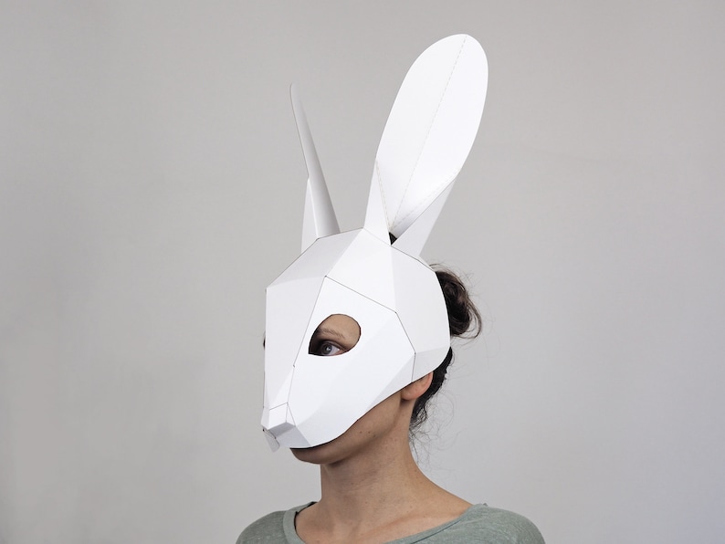 May include: A white geometric paper rabbit mask with long ears and pointed horns. The mask covers the face, with eye holes for visibility. The mask is made of folded paper and has a polygonal design.