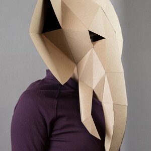 May include: A geometric elephant mask constructed from tan cardboard with a faceted, angular design. The mask features large ears, a long trunk, and triangular eye openings, worn over a solid dark purple long-sleeved shirt.