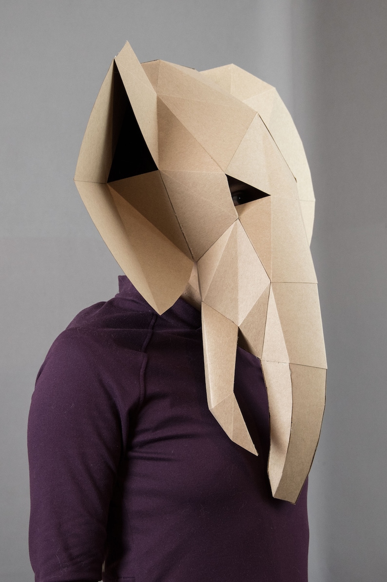 May include: A geometric elephant mask constructed from tan cardboard with a faceted, angular design. The mask features large ears, a long trunk, and triangular eye openings, worn over a solid dark purple long-sleeved shirt.