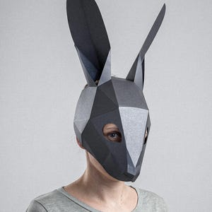 May include: A geometric, low-poly style rabbit mask in shades of grey and black. The mask features large, upright ears and eye openings. The person wearing the mask is wearing a grey t-shirt.