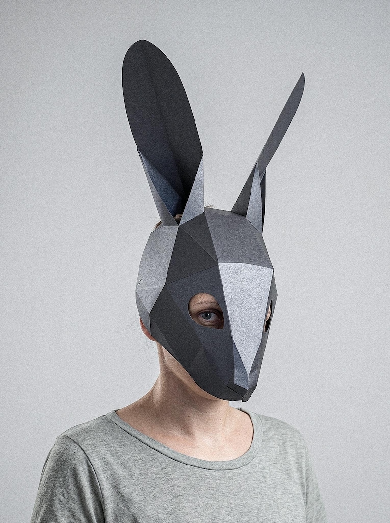 May include: A geometric, low-poly style rabbit mask in shades of grey and black. The mask features large, upright ears and eye openings. The person wearing the mask is wearing a grey t-shirt.
