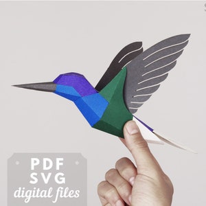 Può includere: Un modello di colibrì in carta artigianale, realizzato con carta blu, verde, viola e bianca. Il modello è tenuto in una mano e ha le ali ripiegate. Il testo "PDF SVG digital files" è in basso nell'immagine.