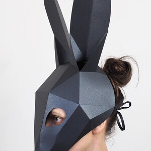 May include: A black geometric rabbit mask with long ears. The mask has a polygonal design and is worn by a person. The mask covers the face, with an opening for one eye. The background is plain white.