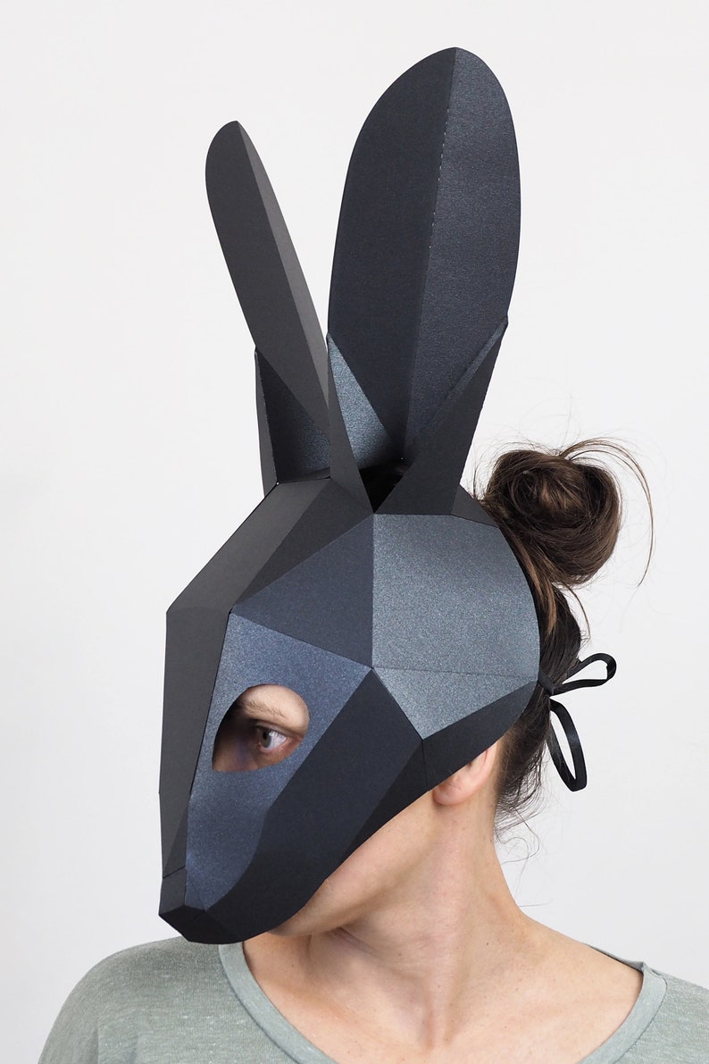 May include: A black geometric rabbit mask with long ears. The mask has a polygonal design and is worn by a person. The mask covers the face, with an opening for one eye. The background is plain white.