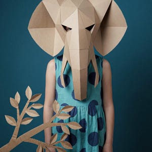 May include: A geometric elephant mask constructed from tan cardboard featuring large ears and a trunk, paired with a separate cardboard branch with leaves. The mask is worn over a sleeveless teal dress with a dark blue polka dot pattern.
