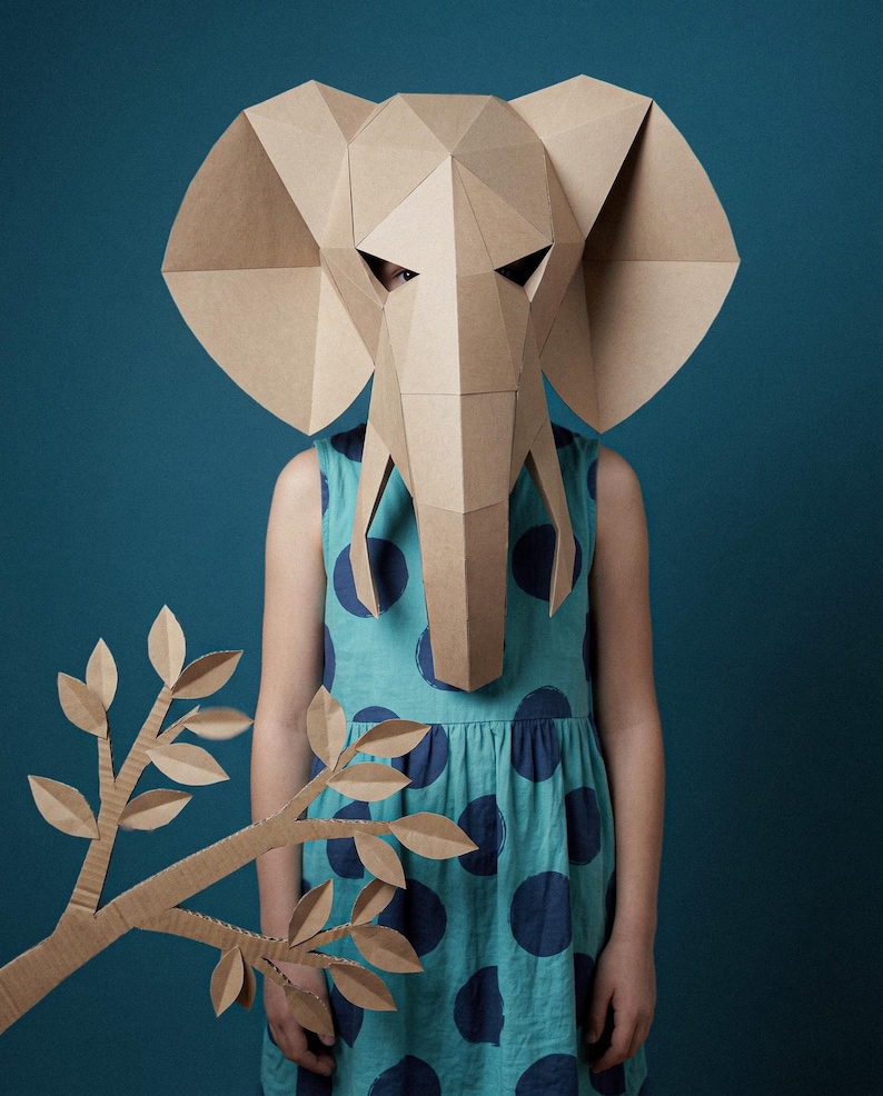 May include: A geometric elephant mask constructed from tan cardboard featuring large ears and a trunk, paired with a separate cardboard branch with leaves. The mask is worn over a sleeveless teal dress with a dark blue polka dot pattern.