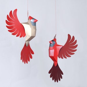 Peut inclure: Deux oiseaux cardinaux en origami rouge et or suspendus &agrave; des ficelles sur un fond blanc. Les oiseaux ont des ailes et des becs d&eacute;taill&eacute;s.