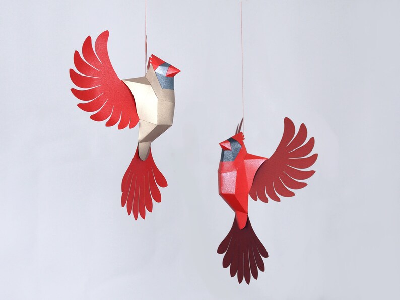 Peut inclure: Deux oiseaux cardinaux en origami rouge et or suspendus &agrave; des ficelles sur un fond blanc. Les oiseaux ont des ailes et des becs d&eacute;taill&eacute;s.