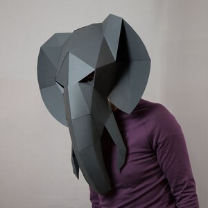 May include: A dark gray, low-poly paper elephant mask featuring a geometric, faceted design with large, flat ears and a long, segmented trunk. The mask covers the face and head, constructed from angular paper panels with visible seams.