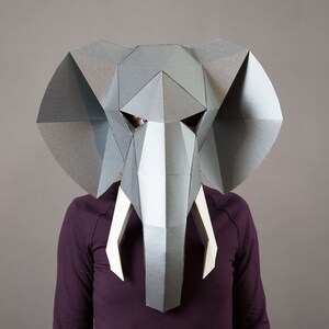 May include: A geometric elephant mask constructed from grey paper panels with large, flat ears and two pointed white tusks. The mask features a faceted, angular design that covers the head and face, showcasing a structured, low-poly aesthetic.