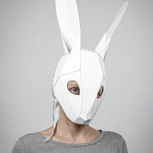 May include: A white geometric rabbit mask with long ears. The mask is made of folded paper and has eye holes. The person is wearing a grey long-sleeve top. The background is a solid grey.