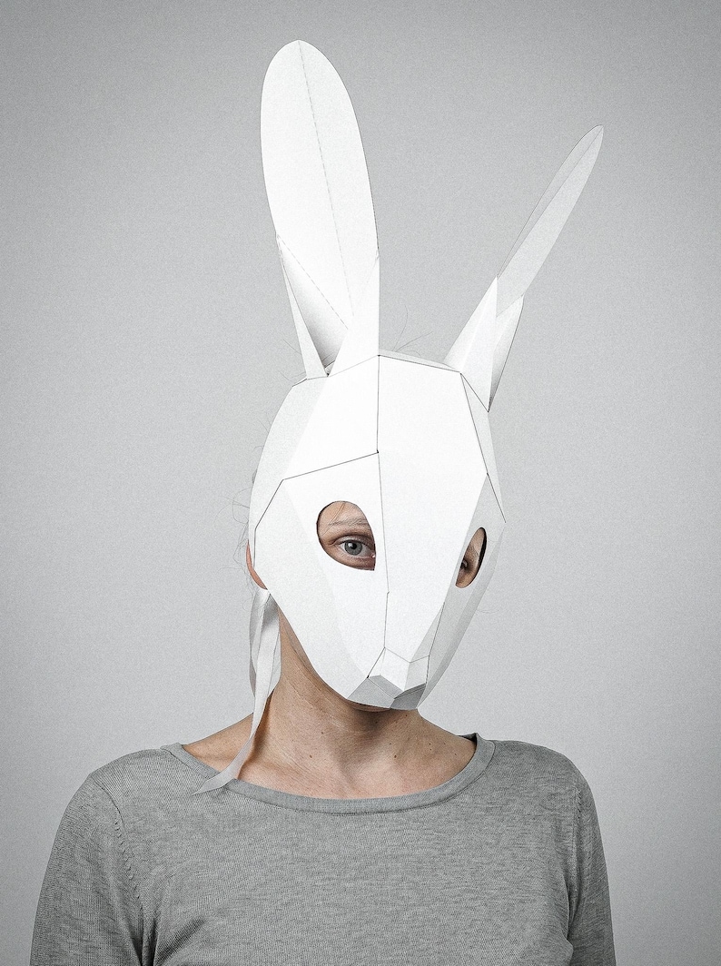 May include: A white geometric rabbit mask with long ears. The mask is made of folded paper and has eye holes. The person is wearing a grey long-sleeve top. The background is a solid grey.