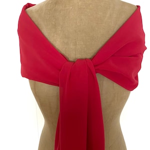 May include: A solid red scarf draped over a beige dress form. The fabric is lightweight and opaque, wrapped around the shoulders and tied in a central knot at the chest, with one end hanging down vertically in a smooth, flowing drape.