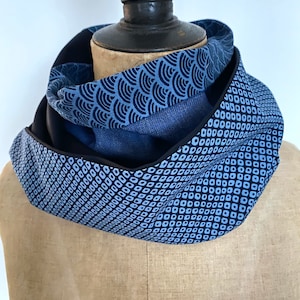 Peut inclure: Écharpe infinie bleue ornée de motifs variés, présentée sur un mannequin. Les motifs comprennent un motif de vagues et un motif géométrique en losanges. L'écharpe est faite d'un tissu doux et possède une bordure noire.