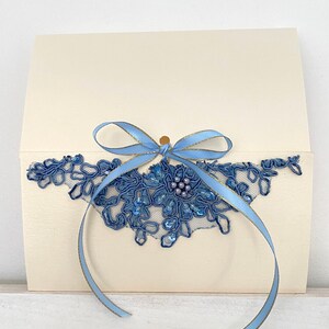Può includere: Biglietto d'invito color avorio con un design in pizzo blu e un nastro azzurro. Il pizzo è impreziosito da paillettes e perline. Il testo "Modèle 1" è in alto.