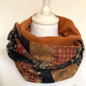 Peut inclure: Écharpe infinie à motifs patchwork. L'intérieur est de couleur rouille, tandis que l'extérieur présente des carrés de tissu noir, rouge et doré avec des motifs floraux et géométriques. Présenté sur une tête de mannequin blanche.