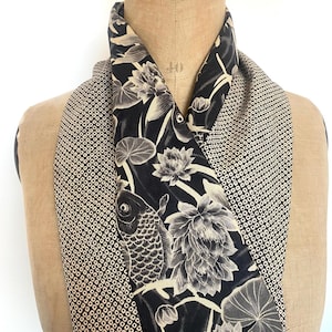 Puede incluir: Bufanda en blanco y negro con un diseño de pez koi y flores de loto. La bufanda está drapeada sobre un maniquí. La tela de fondo tiene un patrón geométrico repetitivo en negro y beige.