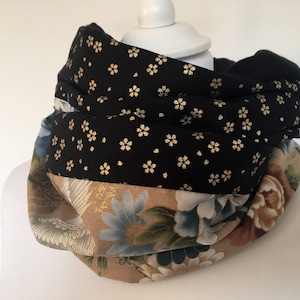 Op de afbeelding: Een meerlagige infinity sjaal met een bloemenpatroon. De bovenste laag is zwart met kleine gouden bloemaccenten. De middelste laag heeft een beige achtergrond met grote blauwe en bruine bloemmotieven. De sjaal is om een witte mannequinhoofd gewikkeld.