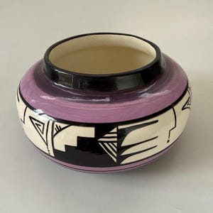 May include: A round ceramic pot with a wide opening. The pot features a lavender band, a black rim, and a cream-colored base with black and white geometric designs. The interior is a light beige color.