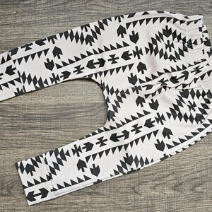 May include: Baby trousers in white with a black geometric pattern. The design includes arrows, triangles, and diamond shapes. The trousers have an elasticated waistband. The fabric appears soft and suitable for comfort.