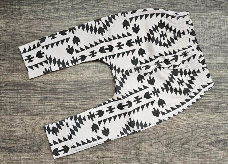 May include: Baby trousers in white with a black geometric pattern. The design includes arrows, triangles, and diamond shapes. The trousers have an elasticated waistband. The fabric appears soft and suitable for comfort.