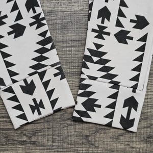 May include: Close-up of two white leggings with a black geometric pattern. The pattern includes arrow, triangle, and diamond shapes. The leggings are folded at the ankles, showing the cuffs.