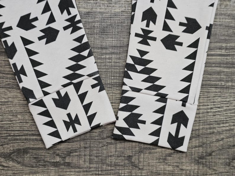 May include: Close-up of two white leggings with a black geometric pattern. The pattern includes arrow, triangle, and diamond shapes. The leggings are folded at the ankles, showing the cuffs.