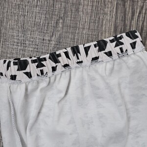 May include: Close-up of white fabric with a black and white geometric patterned waistband. The waistband is elasticated and sewn to the white fabric. The fabric appears to be lightweight, possibly part of a garment.