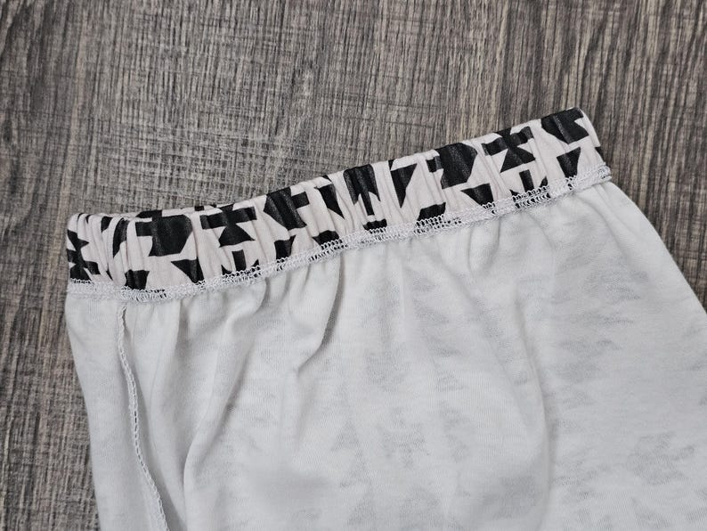May include: Close-up of white fabric with a black and white geometric patterned waistband. The waistband is elasticated and sewn to the white fabric. The fabric appears to be lightweight, possibly part of a garment.