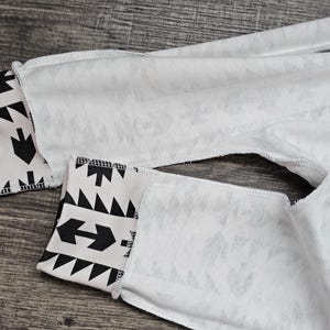 May include: Close-up of white fabric with black geometric patterns on the cuffs. The cuffs feature arrows, triangles, and other shapes. The fabric appears to be a light material, possibly cotton or a blend.