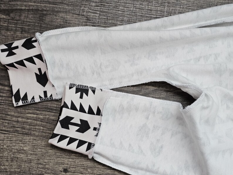 May include: Close-up of white fabric with black geometric patterns on the cuffs. The cuffs feature arrows, triangles, and other shapes. The fabric appears to be a light material, possibly cotton or a blend.