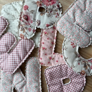 May include: A selection of fabric letters in assorted patterns. Some letters display floral designs in pink and cream, while others have a pink and white gingham pattern. The letters have scalloped edges.