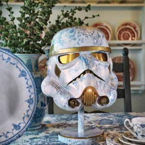 May include: A Stormtrooper helmet display piece featuring a white and blue floral toile pattern. The helmet includes gold-colored accents on the brow, eye lenses, and mouth area, and it is mounted on a matching circular stand with a floral finish.
