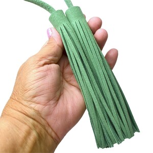 May include: Two sage green suede leather tassels featuring long, thin, vertical fringe strips attached to a cylindrical cap with matching green leather cords extending from the top, presented against a plain white background.