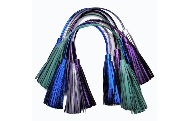May include: A collection of six decorative tassels featuring metallic finishes in shades of blue, purple, teal, and silver. Each tassel is attached to a long, thin cord, creating a fan-like arrangement of hanging fringe elements with a reflective texture.