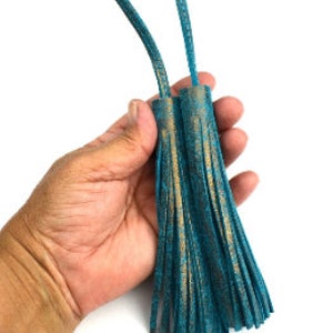 May include: Two turquoise-colored tassels with bronze accents. The tassels are made of a soft, suede-like material and are attached to a thin cord. The tassels are approximately 5 or 6 inches long.