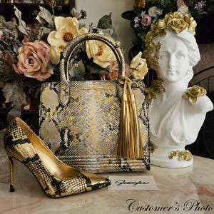 May include: A structured handbag and a matching high-heeled pump featuring a gold, black, and grey snakeskin pattern. The handbag includes two top handles and a gold tassel charm. A white card with the handwritten name Jennifer is placed nearby.