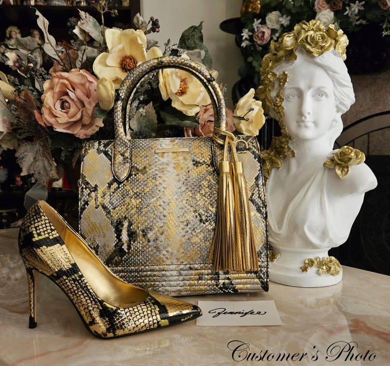 May include: A structured handbag and a matching high-heeled pump featuring a gold, black, and grey snakeskin pattern. The handbag includes two top handles and a gold tassel charm. A white card with the handwritten name Jennifer is placed nearby.