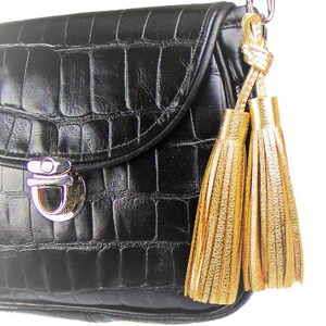 May include: A black handbag with a textured crocodile-embossed pattern, featuring a silver-toned metal push-lock closure on the front flap. Two metallic gold tassels are attached to the side of the bag by a silver-toned metal lobster clasp.
