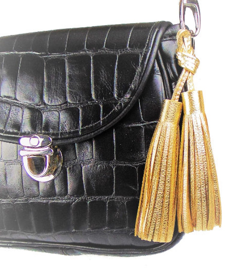 May include: A black handbag with a textured crocodile-embossed pattern, featuring a silver-toned metal push-lock closure on the front flap. Two metallic gold tassels are attached to the side of the bag by a silver-toned metal lobster clasp.
