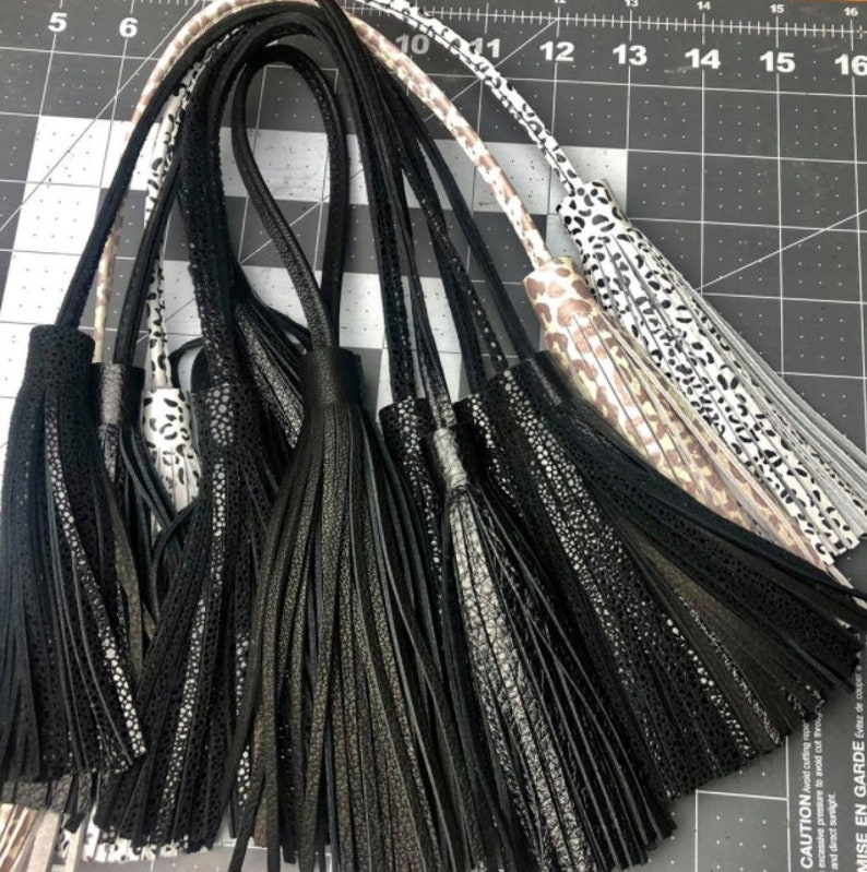 May include: A collection of hanging tassels featuring black, white, and tan colors with various textures including solid black, speckled, and leopard print patterns. Each tassel is attached to a long, thin cord, displayed against a cutting mat.